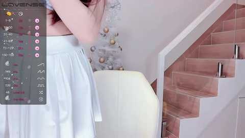 Video of a woman in a white dress, facing away, standing near a beige armchair in a modern, minimalist room with a pink-tiled staircase and white walls.