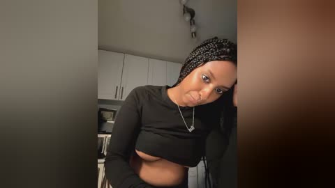 Media: Video of a young Black woman with braided hair, wearing a black crop top and necklace, leaning against a kitchen counter.