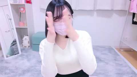 Media: Video of an East Asian woman with long black hair, wearing a white mask, sitting on a grey carpeted floor in a brightly lit room. She's dressed in a white long-sleeved top and black pants, with a pink lamp and white shelves in the background.