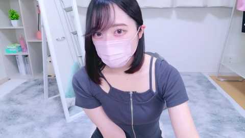 Media: Video of a young Asian woman with straight black hair, wearing a gray top, pink mask, and glasses, kneeling on a gray carpeted floor in a bright, minimalist room with white walls and a mirror.