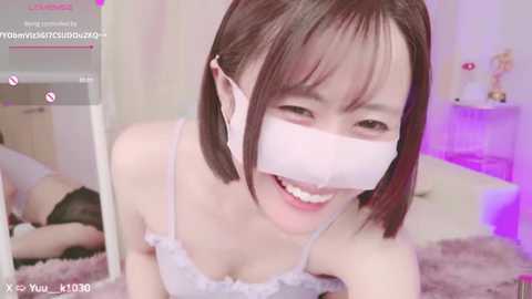 Media: Video of a smiling Asian woman with short brown hair, wearing a white face mask, pink bra, and black panties, lying on a bed with a purple blanket, in a cozy bedroom.