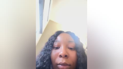 Media: Video of a young Black woman with medium brown skin and curly hair, wearing a neutral expression. The background features a light beige wall with a window frame, suggesting an indoor setting.