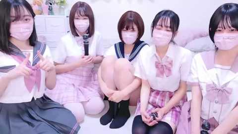 Media: Video of five Japanese schoolgirls in matching sailor uniforms, pink plaid skirts, and face masks, sitting on a white floor, holding hands in a circle, in a bright, clean room.