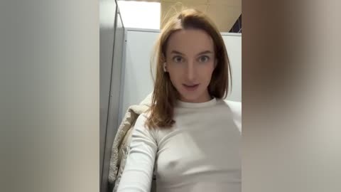 Media: A video of a fair-skinned woman with long, straight auburn hair, wearing a white long-sleeve top, sitting in a white-walled bathroom. She has a neutral expression, and the background includes a beige towel and a white door.