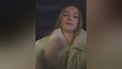 Media: Video of a young Caucasian woman with fair skin and light brown hair, wearing a beige fur coat, standing indoors in a dimly lit room.