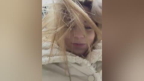 Media: Video of a blonde woman with messy hair, partially obscured by her bangs, wearing a white fluffy jacket, with a blurred background.