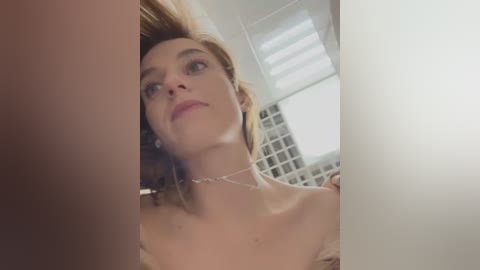 Media: Video of a fair-skinned woman with light brown hair, wearing a silver necklace, standing in a modern bathroom with white tiles and a large window. She looks upward, expression neutral.