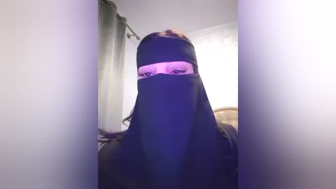 Media: A video of a woman with long dark hair wearing a black niqab, her eyes visible through a small slit, in a dimly lit room with white walls and a window covered by a green curtain.