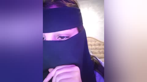 Media: Video of a woman with light skin and brown hair, partially obscured by a black veil, holding a finger to her lips in a bedroom with a beige quilted headboard.