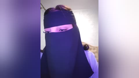 Media: A video of a person wearing a black niqab with a slit for the eyes, partially visible, in a dimly lit room with a beige couch and a white wall.