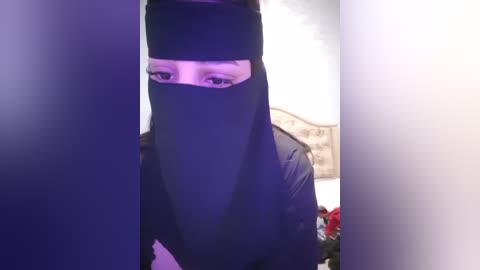 Media: Video of a person wearing a black niqab, obscuring their face except for their eyes. Background includes a beige headboard and blurred, indistinct objects.