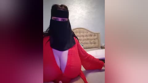 Media: Video of a woman with long, dark hair wearing a black veil over her face, a red blouse, and a white shirt underneath. She sits at a beige upholstered headboard in a dimly lit room.