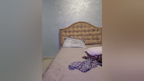 Media: A video of a small, cluttered bedroom with a light blue wall and a beige upholstered headboard. The bed has a wrinkled beige sheet, a purple and white patterned pillow, and a disheveled pile of clothes on the floor.