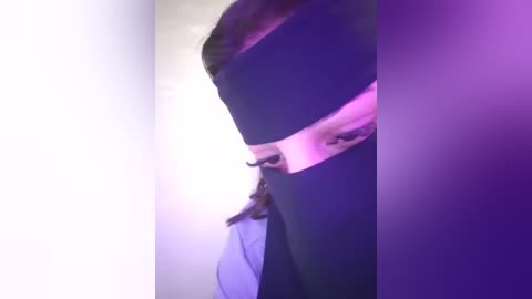 Media: A video captures a close-up of a person's face, partially obscured by a black blindfold. The subject's head is turned slightly to the left, and the background is illuminated with a soft, purple hue, suggesting a dimly lit room. The person's long, brown hair is visible.