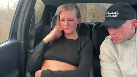 Media: Video of a blonde woman in a black crop top, seated in a car, looking tired. A man in a baseball cap and light jacket sits beside her.