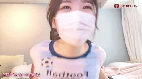 Media: A blurry video of an Asian woman with fair skin wearing a white mask and a white shirt with \"Madool\" text, taken indoors in a bedroom with white walls and a bed.