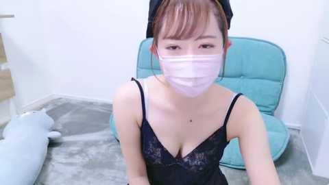 Media: Video of an Asian woman with light skin, wearing a black lace camisole, blue headband, and a pink face mask, kneeling on a gray carpet in a room with a teal chair and plush toys.