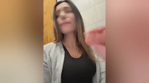 Media: A blurry video of a smiling woman with long brown hair, wearing a black top and grey cardigan, in a bathroom with white tiles and pink towels.