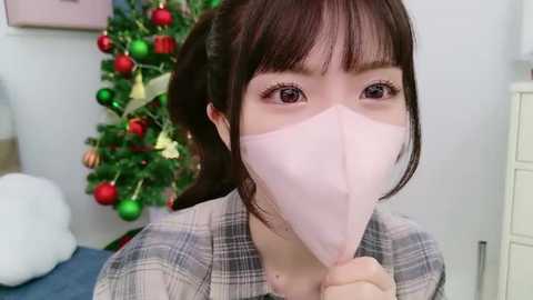 Media: Video of an Asian woman with straight brown hair and bangs, wearing a pink face mask, grey plaid shirt, and a white sweater, standing in front of a decorated Christmas tree with red and green ornaments.