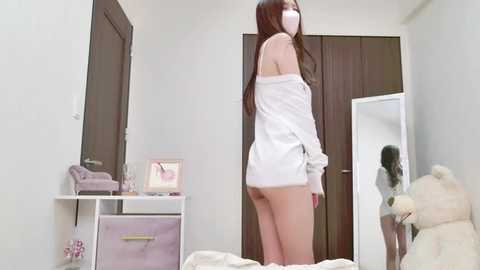 Media: Video of an East Asian woman in white off-shoulder top, revealing her bare buttocks, standing in a minimalist bedroom with a mirror, stuffed toys, and pastel-colored storage.