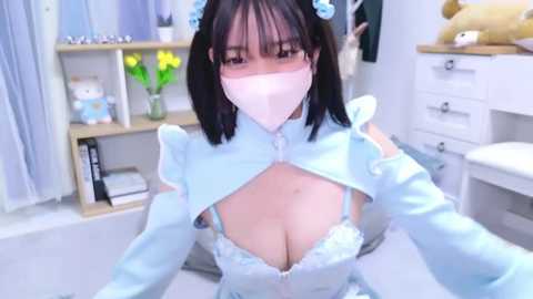 Media: A video of an East Asian woman with black hair, wearing a light blue maid cosplay outfit, featuring a low-cut bodice revealing her large breasts. She has a pink mask covering her face. The background shows a white room with shelves and stuffed toys.