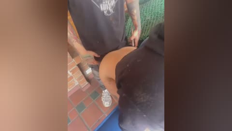 Media: Video of a man with a tattooed arm, wearing a black T-shirt, bending over to expose his bare buttocks. Background shows a red-tiled floor and a green netted wall.