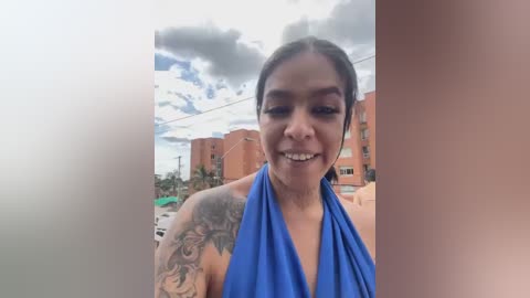 Media: Video of a smiling Latina woman with medium skin tone, dark hair tied back, wearing a blue halter top, showcasing a large tattoo on her left arm. Background features a cloudy sky, red-brick buildings, and palm trees.