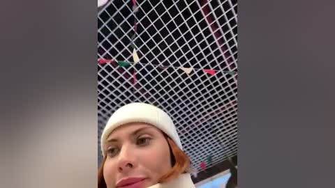 A video of a woman with light skin and auburn hair wearing a white beanie and a light jacket, standing in front of a black-and-white diamond-patterned chain-link fence.