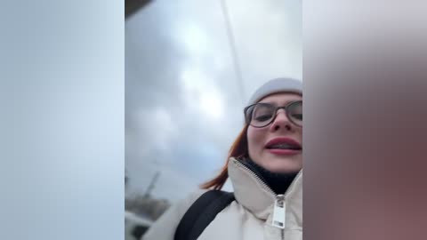 Media: Video of a woman with red hair, wearing glasses, a light jacket, and a white beanie, smiling slightly, standing in front of a reflective surface, creating a distorted, mirrored effect.