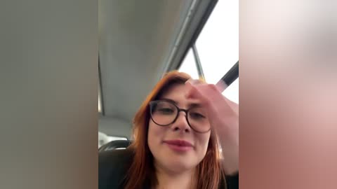 Media: A video shows a young woman with red hair, wearing black-framed glasses, smiling inside a car. The background reveals a blurred window and interior details.