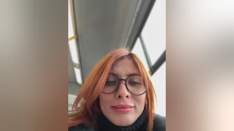 Media: Video of a fair-skinned, red-haired woman with glasses, wearing a black turtleneck sweater, standing indoors next to a large window with a white frame.