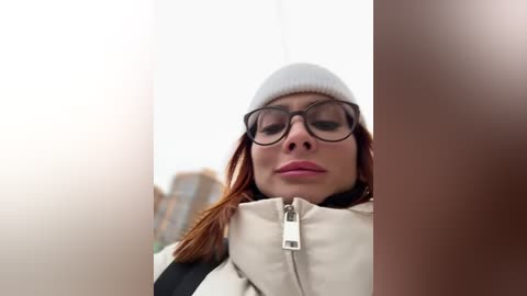 Video of a young woman with fair skin, red hair, and large, dark-framed glasses. She wears a white beanie, beige jacket, and carries a black backpack. The background is blurred, featuring a cityscape with tall buildings.