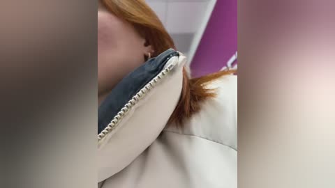 Media: Video of a woman with red hair lying on a white leather couch, wearing a blue denim jacket, partially covered by a beige blanket, against a purple wall background.