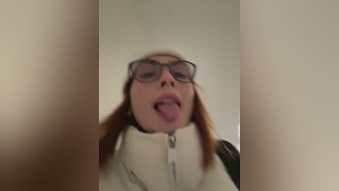 Media: Video of a young woman with long, straight red hair, wearing large, dark-rimmed glasses, sticking out her tongue, and zipped up in a white jacket. The background is a plain, beige wall, emphasizing her playful, slightly mischievous expression.