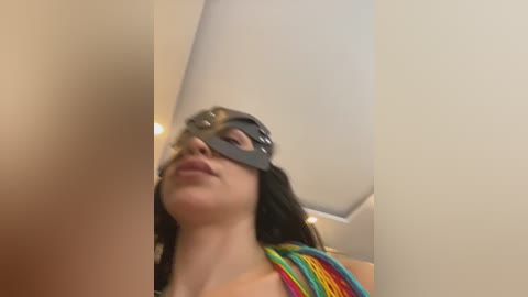 Media: A video of a woman with long brown hair, wearing a black blindfold, standing in a room with a white ceiling and a colorful rainbow-striped towel.