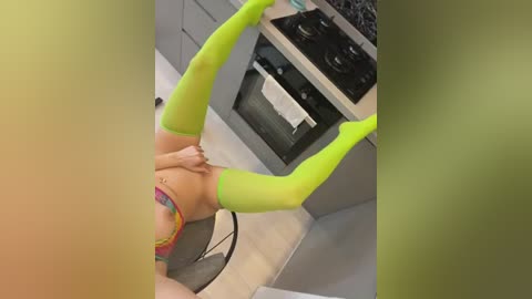 Media: A video captures a woman with light skin, wearing neon green thigh-high stockings and a colorful headband, performing a headstand in a modern kitchen.