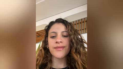 Media: Video of a young woman with curly, wavy brown hair, light skin, and closed eyes, smiling slightly. She wears a black top. The background features wooden blinds and a beige wall.