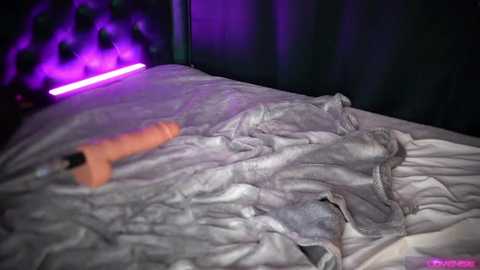 Media: A video of a messy bedroom with a rumpled gray blanket, an orange vibrator on the bed, and a purple neon light emitting a soft glow.