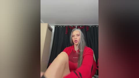 Media: Video of a woman with long, straight, silver hair, wearing a red sweater, seated on a bed with black curtains. She has a slightly open mouth and looks towards the camera.