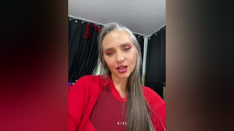 Media: A video of a woman with long, straight, silver-gray hair, wearing red lipstick and eye makeup, in a red top, standing in a dimly lit room with black curtains.