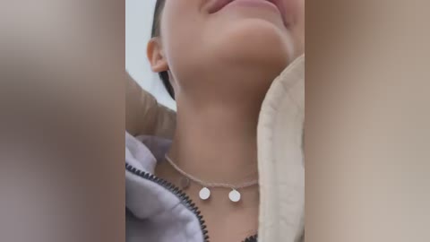 Media: Video of a close-up of a woman with light skin, wearing a silver necklace and a grey zip-up jacket, smiling.