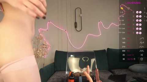 Media: Video of a woman in pink lingerie with a small breast size, standing in a room with a green couch, neon lights, and a glass table.