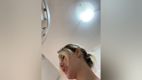 Media: A video of a blonde woman with a ponytail, wearing a top, looking upward, in a white room with a circular ceiling light and clean walls.