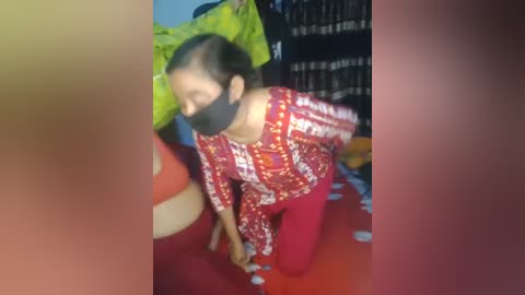 Media: Video of a woman in a red-patterned blouse and red pants kneeling on a red bedspread, wearing a black mask, in a dimly lit room with a green and black patterned wall and a blue wall.