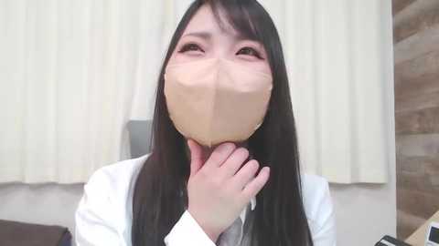 Media: Video of a young Asian woman with long black hair, wearing a white mask, white shirt, and a white jacket, holding her chin, in a room with beige curtains and a wooden wall.