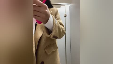 Media: Video of a person in a beige coat, holding a pink object, standing in a bathroom with white walls and a door.