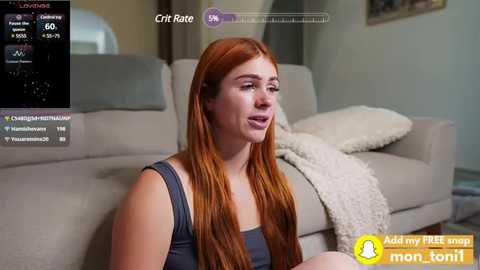 Media: Video of a red-haired woman in a gray tank top, sitting on a beige couch, with a white blanket draped over the armrest.