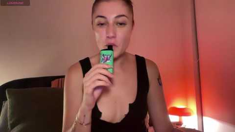 Media: Video of a light-skinned woman with brown hair in a ponytail, wearing a black tank top, vaping in a dimly lit bedroom with a red lamp and bed.