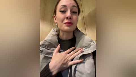 Media: A video of a fair-skinned woman with straight brown hair, wearing a light gray puffer jacket over a black shirt, posing indoors with her hands clasped over her chest, giving a serious expression.