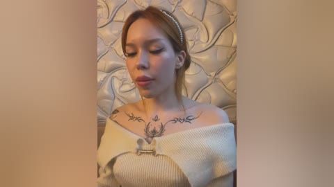 Media: Video of a young woman with light skin and brown hair, wearing a cream off-shoulder sweater, adorned with a necklace and tribal tattoo on her chest, lying on a tufted headboard.